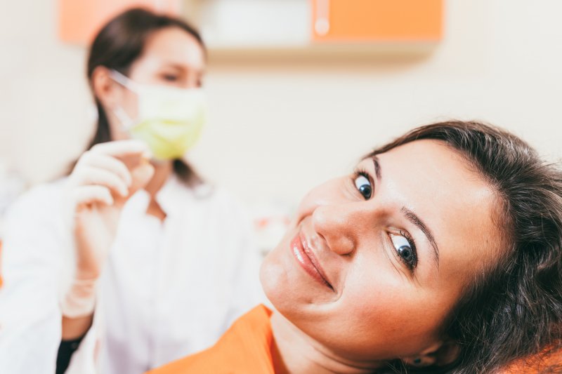 a young woman lying in a dentist’s chair smiling while her dentist holds an extracted tooth in the background